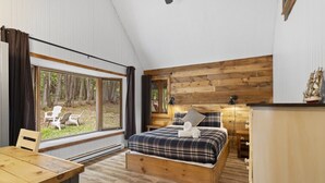 Un dormitorio con cama, cabecera de madera, ventana con vista a árboles y una mesa con sillas.