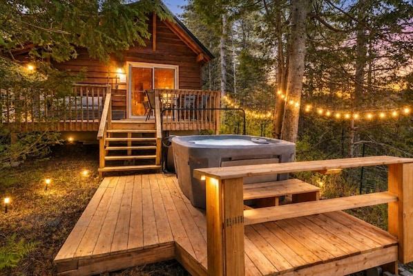 A wooden cabin with a hot tub, wooden deck, and string lights.