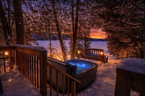 A hot tub surrounded by snow-covered deck railings, with a view of a lake and trees at dusk.
