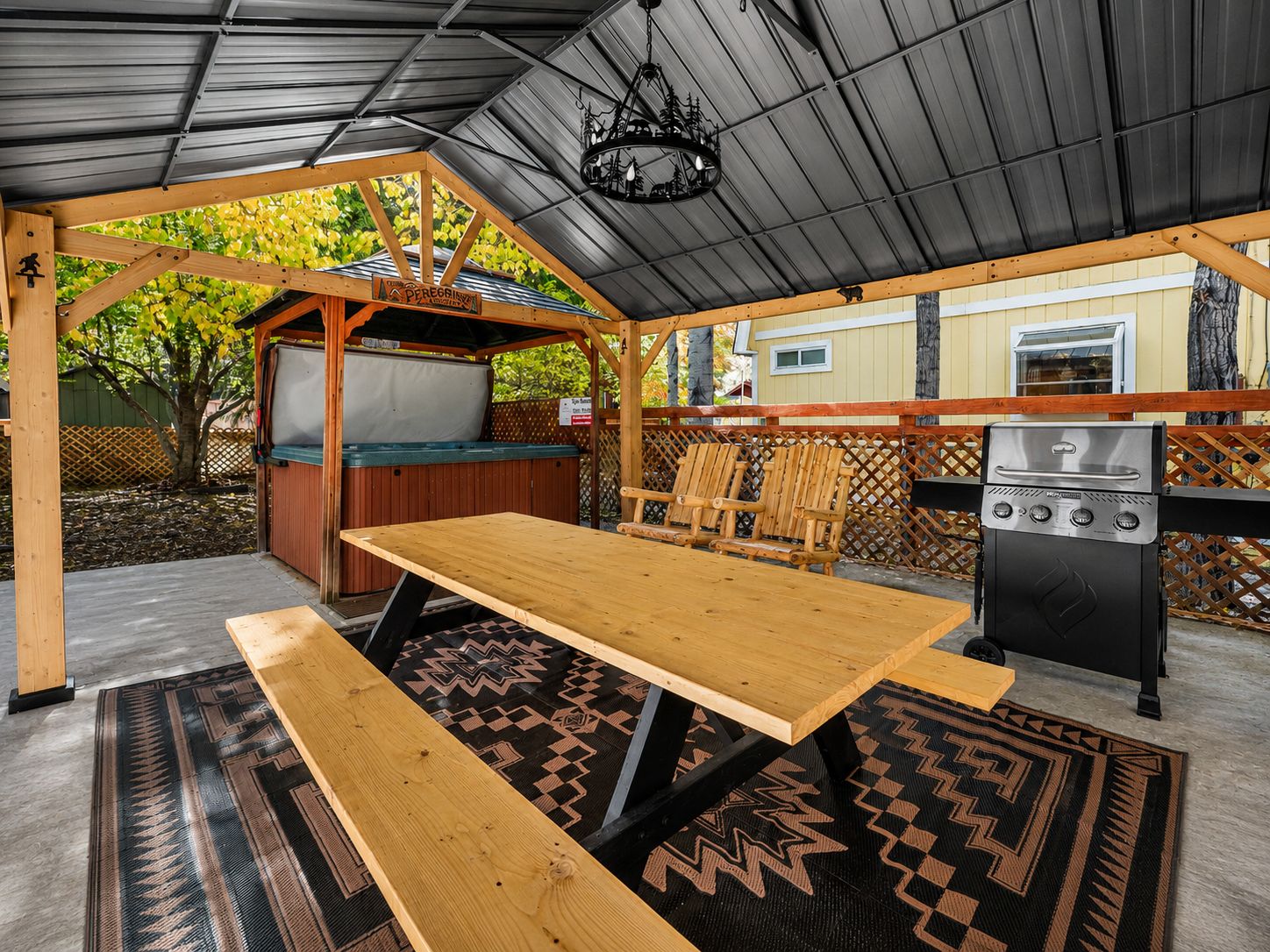 Un patio exterior con mesa de madera, bancos, una parrilla y un área de estar techada.