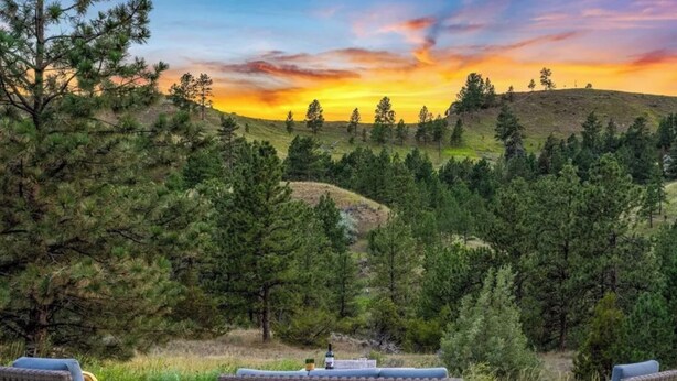 A scenic outdoor seating area with wicker furniture and a blanket, overlooking a forested hillside. - Montana Retreat (Billings)