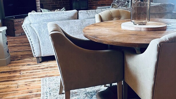 A cozy living room with a round wooden table, beige leather chairs, a grey sofa, and a brick fireplace. - Farnah House Barn (Belper)