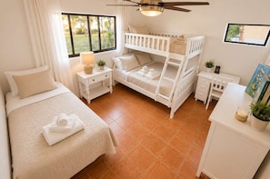 A bedroom with a bunk bed, a single bed, a desk, and a window with a view of greenery.