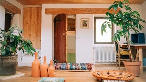 Ein gemütliches Zimmer mit Holzbalkendecke, Holztür, Fenster, Heizkörper, Bücherregal und einer großen Pflanze.