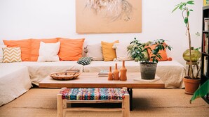 Ein gemütliches Wohnzimmer mit einer Couch, Kissen, einem Couchtisch, einer kleinen Bank, einer Topfpflanze und einem Bücherregal.