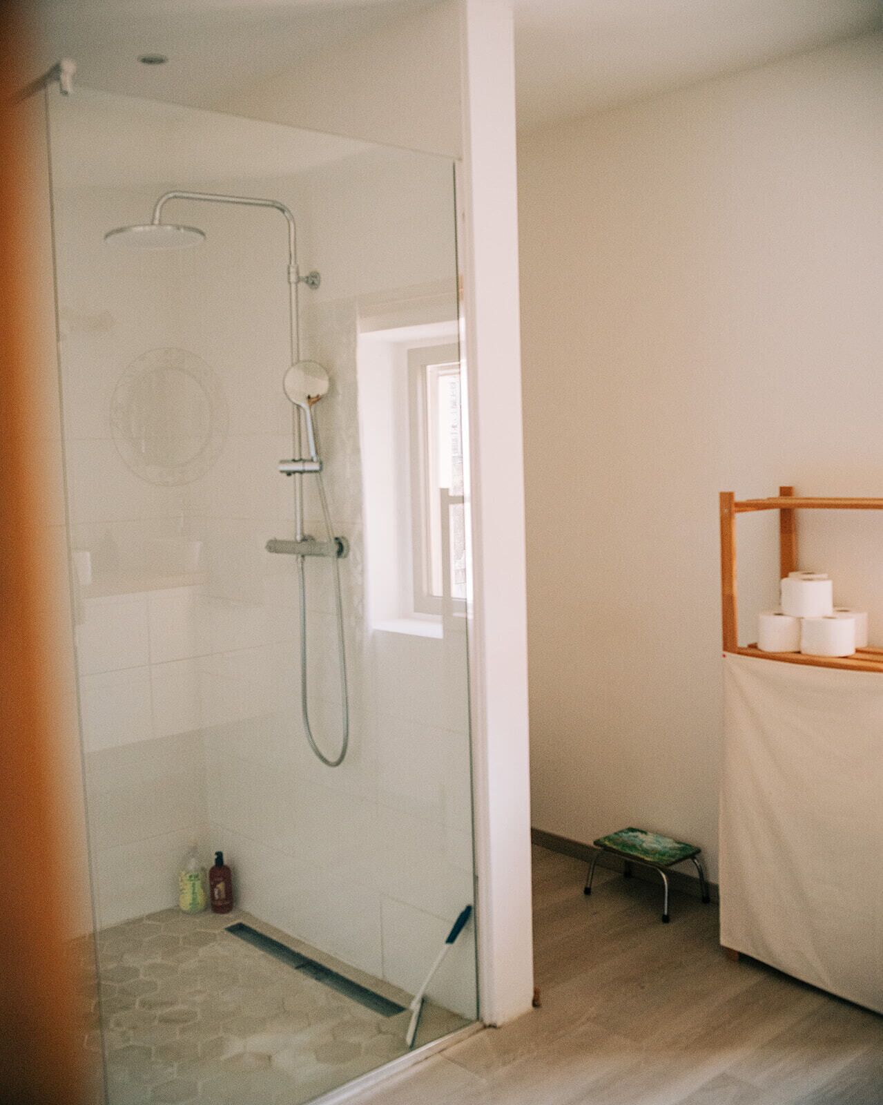 Une salle de bain avec une douche à parois de verre, un petit marchepied et une étagère avec du papier toilette.