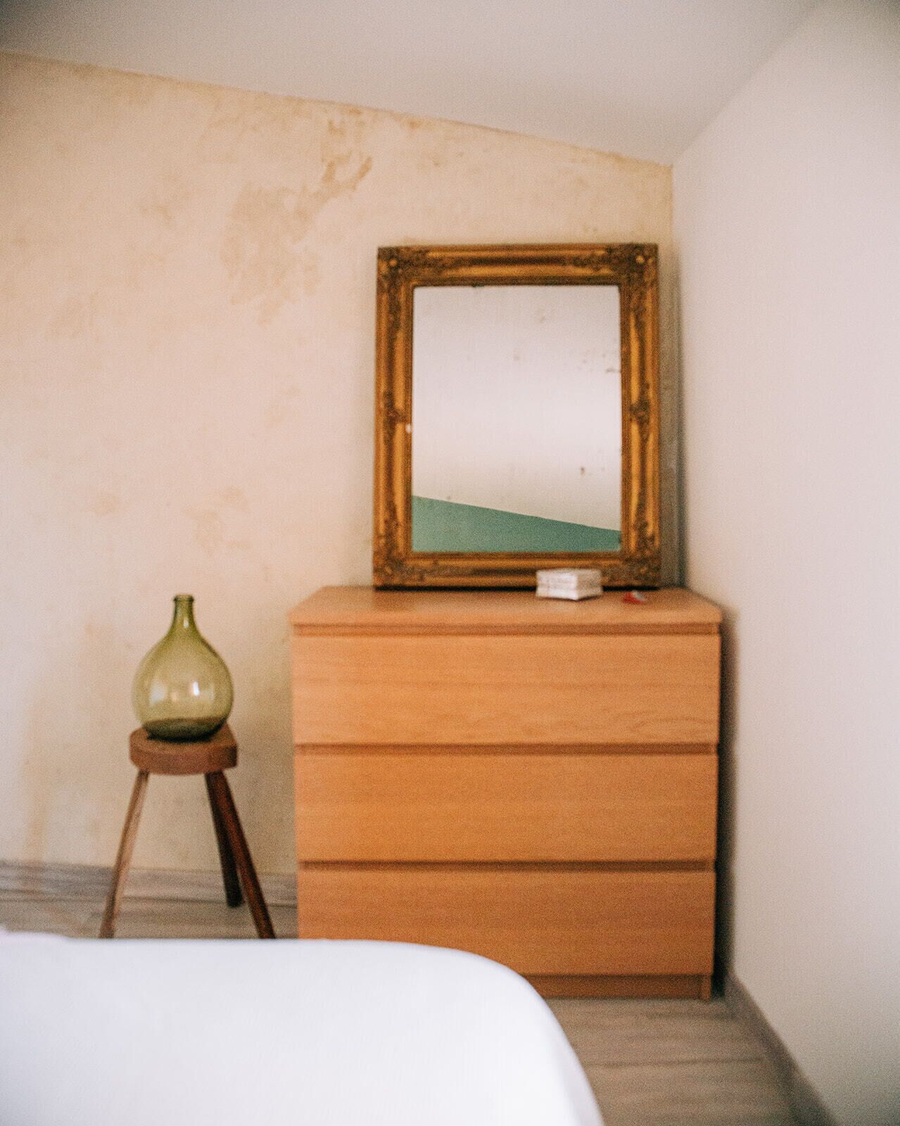 A room with a wooden dresser, a mirror with a wooden frame, and a small green vase on a stool.