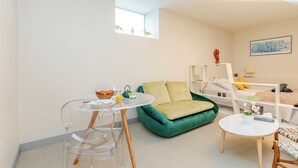A modern room with a green sofa, a white bed with a headboard, a small round table with a chair, and a window with a view of the sky. - Spacious studio, chic and quiet, attractive Bon Marché district (Paris)