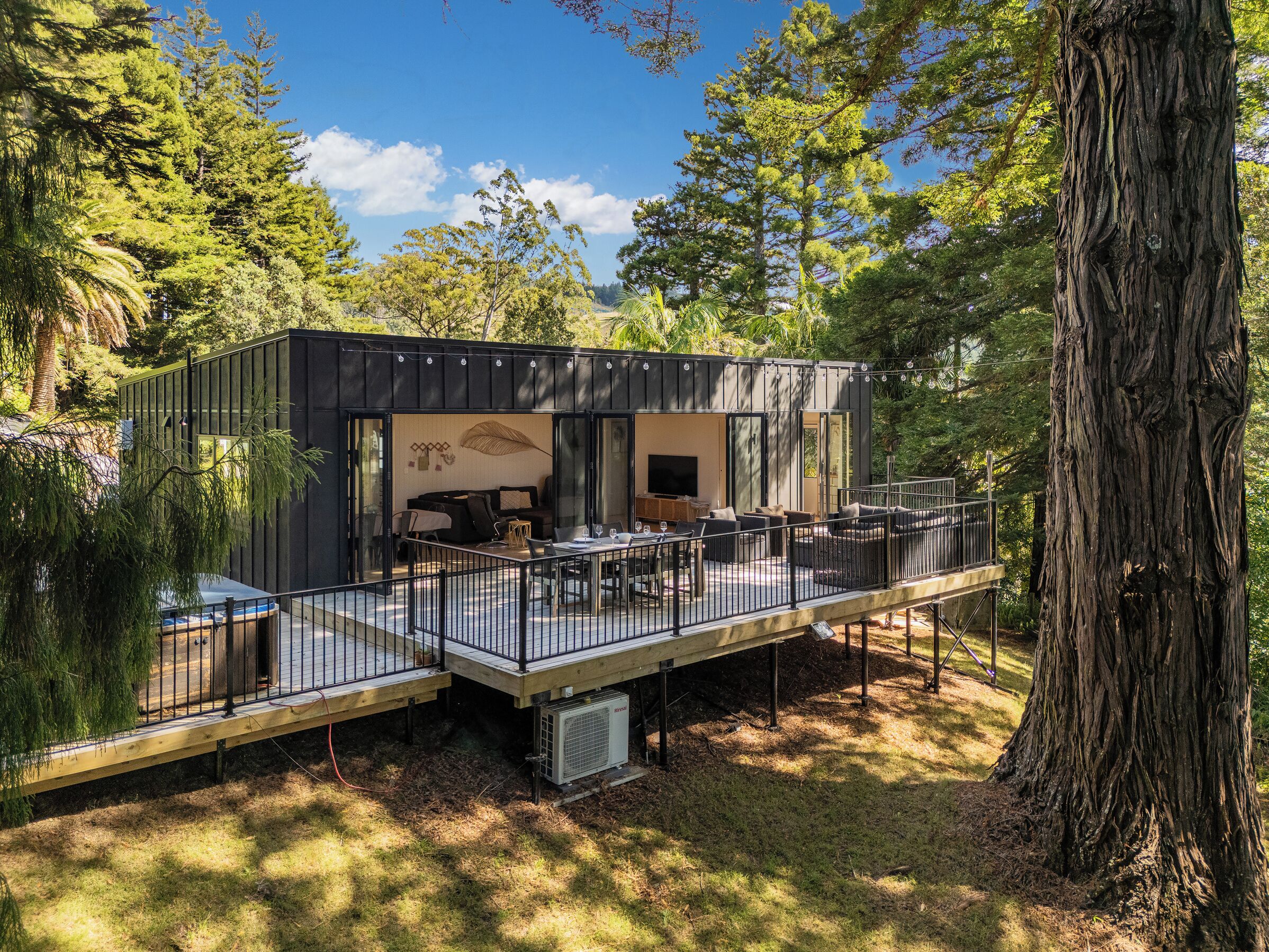 Een moderne cabin met terras, buitenmeubels en een grote boom in de buurt.
