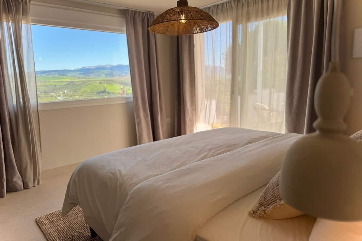 Ein Schlafzimmer mit einem großen Bett, einer hängenden Leuchte und Blick auf eine grüne Landschaft durch das Fenster.