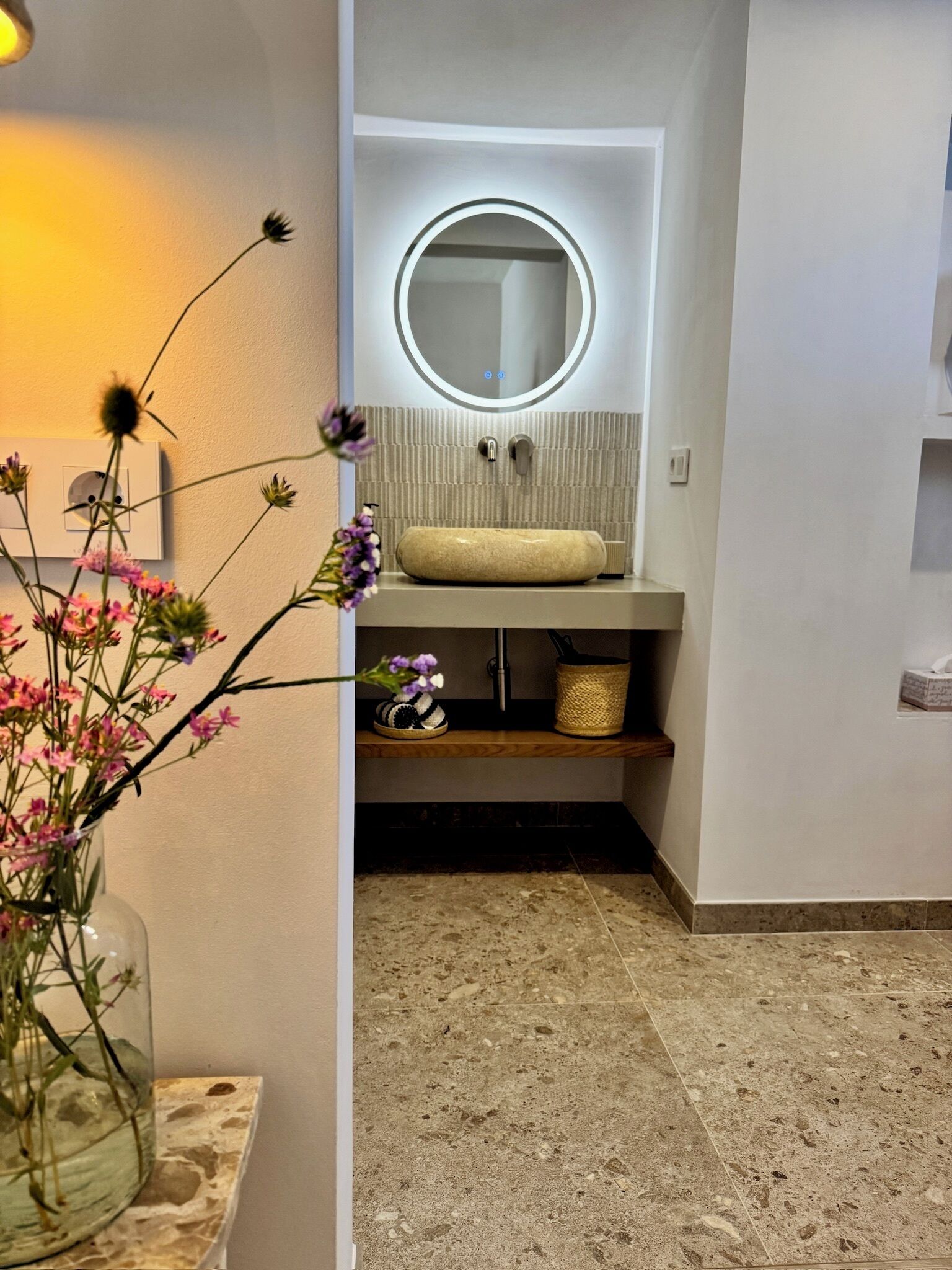 Un baño con un espejo redondo, un lavabo color beige, una pequeña estantería con una cesta y un florero con flores.