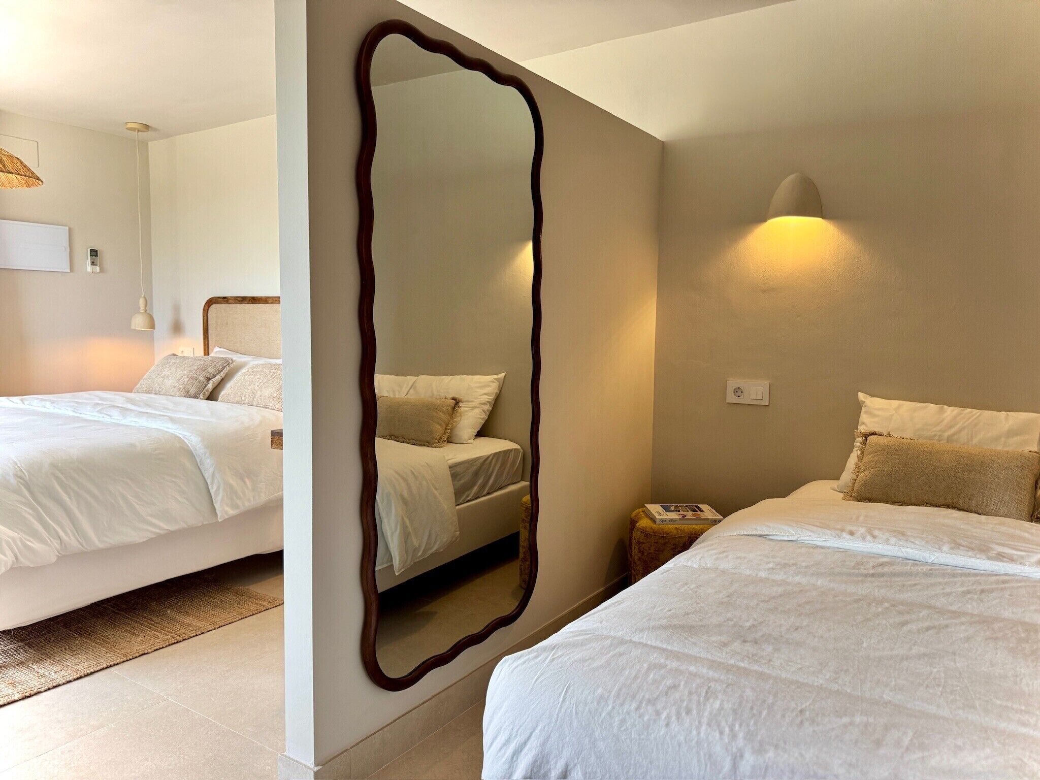 A hotel room with two beds, a large mirror, a bedside table, a lamp, and a wall-mounted light fixture.