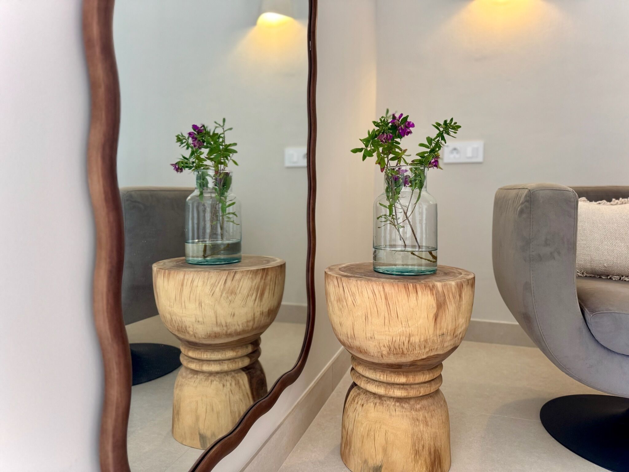 A room with a wooden side table, a grey sofa, and a mirror reflecting the room.