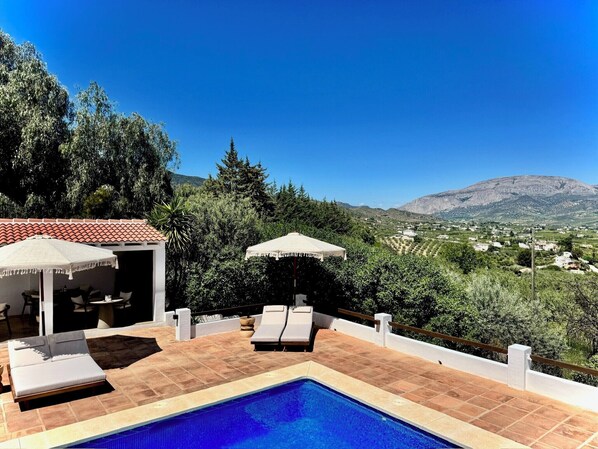 Un espace piscine doté de chaises longues et de parasols, entouré de verdure et avec une chaîne de montagnes en toile de fond.