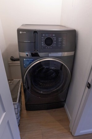 A compact, modern washer and dryer unit with a control panel, located in a small laundry room with a wooden floor and a striped laundry basket. - Lovely 1-bedroom  guest house with AC in tranquil Tucson. (Tucson)