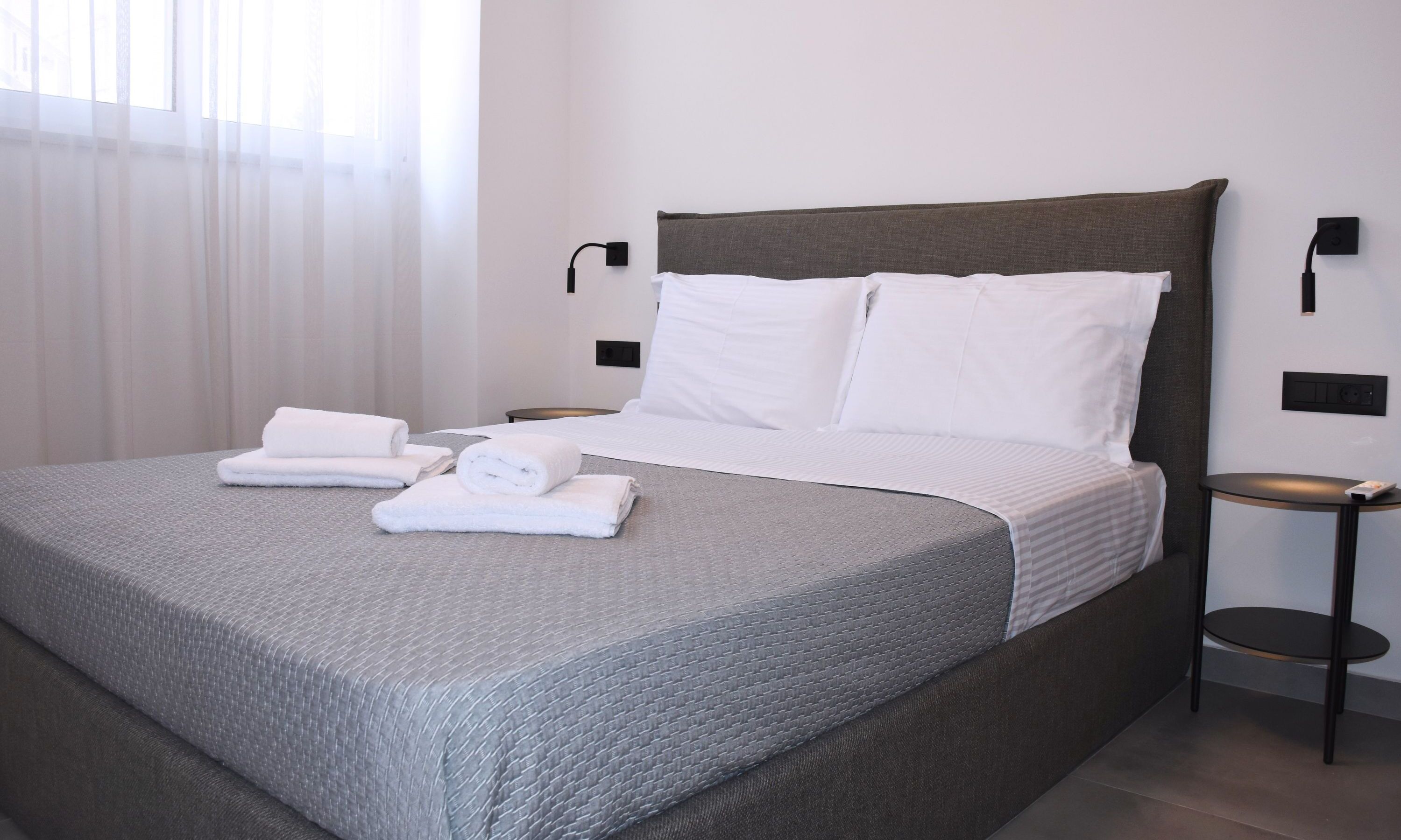A neatly made bed with white linens and towels, a grey textured bedspread, and a dark headboard.