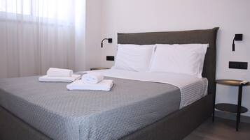 A neatly made bed with white linens and towels, a grey textured bedspread, and a dark headboard.