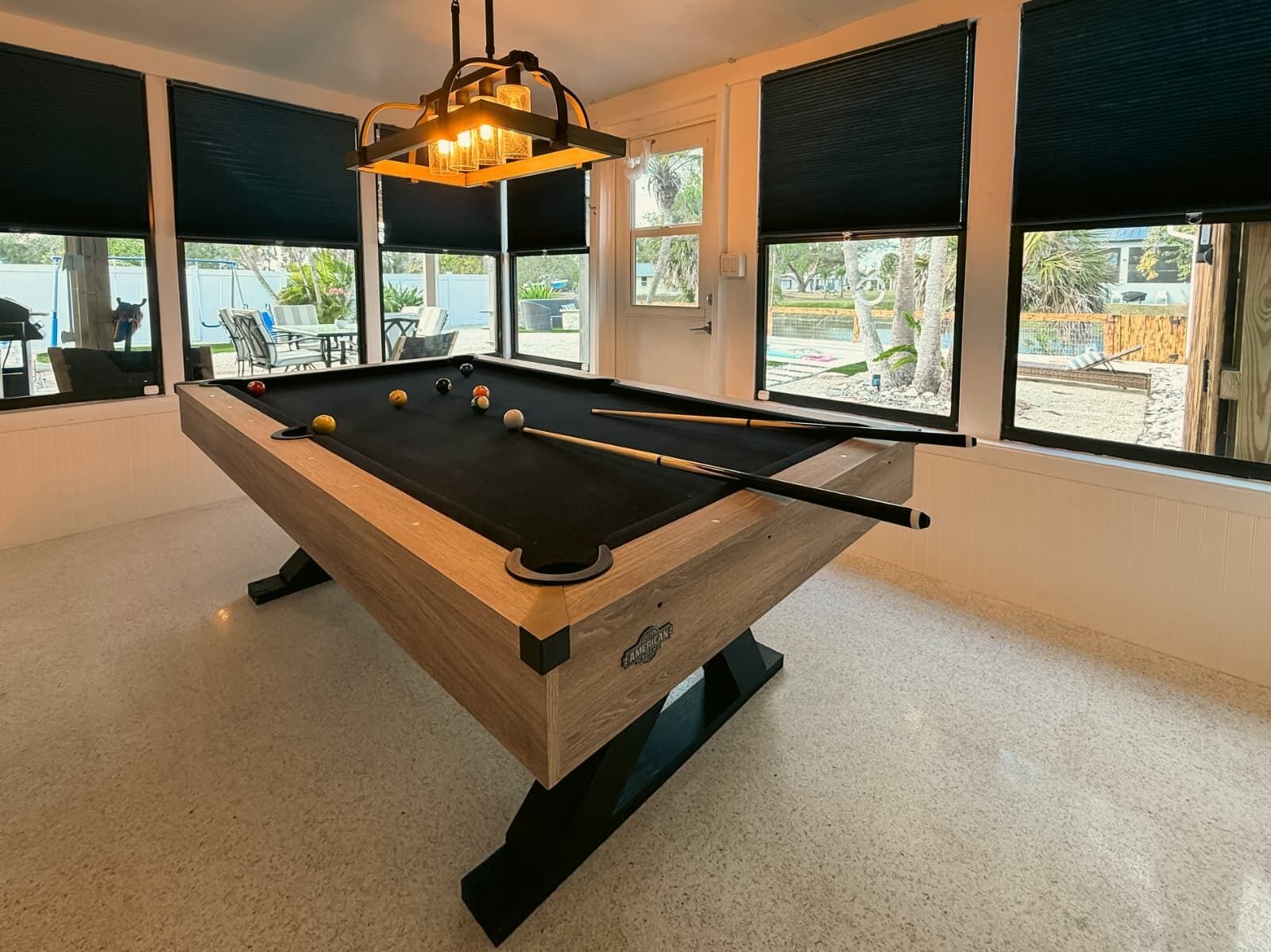 Uma mesa de bilhar com acabamento em madeira e superfície em feltro preto, localizada em um cômodo com janelas amplas e um lustre.