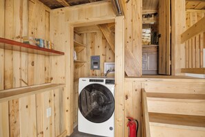 A compact wooden room with a washing machine, a shelf with books, and a fire extinguisher.