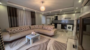 A modern living room with a sectional sofa, a coffee table, and a kitchen in the background. - LOVELY  RESİDENCE 2+1 CLOSE TO SEA WİTH POOL (mahmutlar)