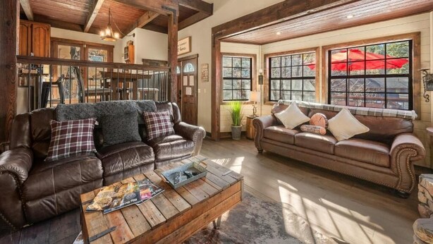Una sala rústica con sofás de cuero, vigas de madera y ventanas amplias con sombrillas rojas en el exterior.