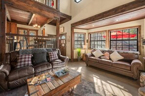 A rustic living room with leather sofas, wooden beams, and large windows with red umbrellas outside.