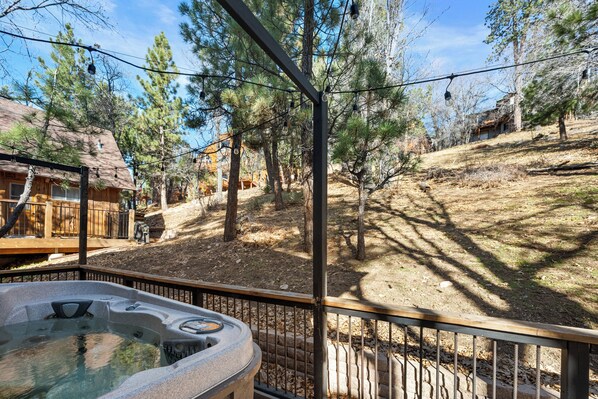 A hot tub area with a wooden deck and a fenced enclosure, surrounded by trees and a hilly landscape.