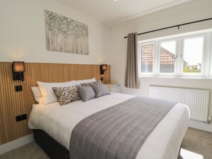 Ein Schlafzimmer mit Bett, Kopfteil, Wandbildern, Nachttischlampen, einem Fenster mit Vorhängen und einem Heizkörper.