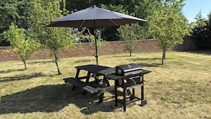 Ein Picknickplatz mit Tisch, Bank, Grill und Sonnenschirm in einer grünen Umgebung.