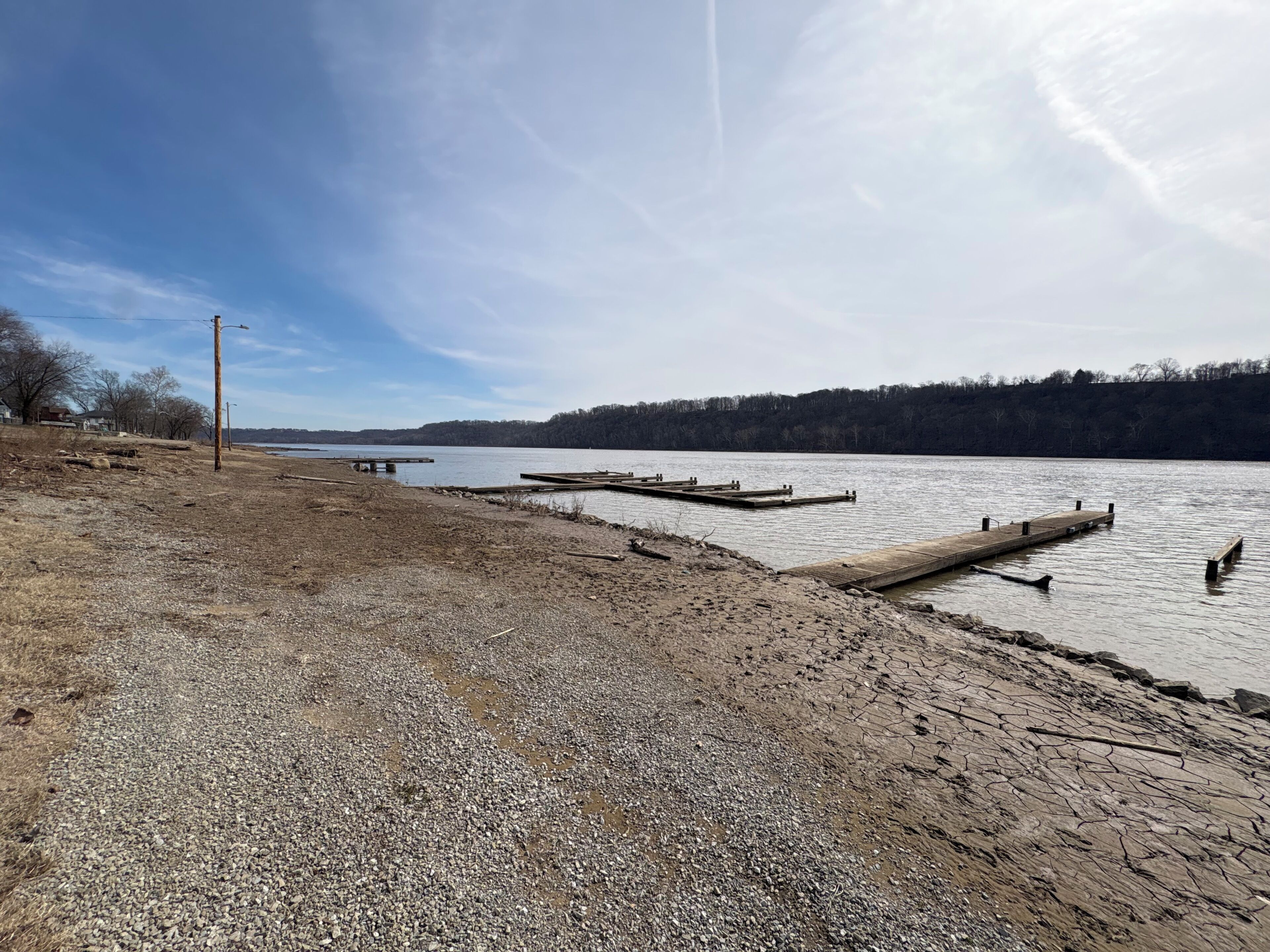 A gravel beach with wooden docks extending into a calm lake, surrounded by trees and a clear sky.