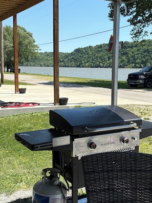 Una parrilla Blackstone con tanque de propano, instalada al aire libre con vista a un lago y árboles.