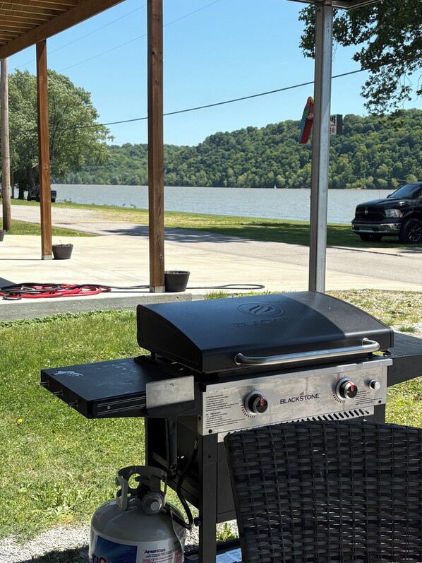 Un barbecue Blackstone avec une bouteille de propane, installé à l’extérieur, offrant une vue sur un lac et des arbres.