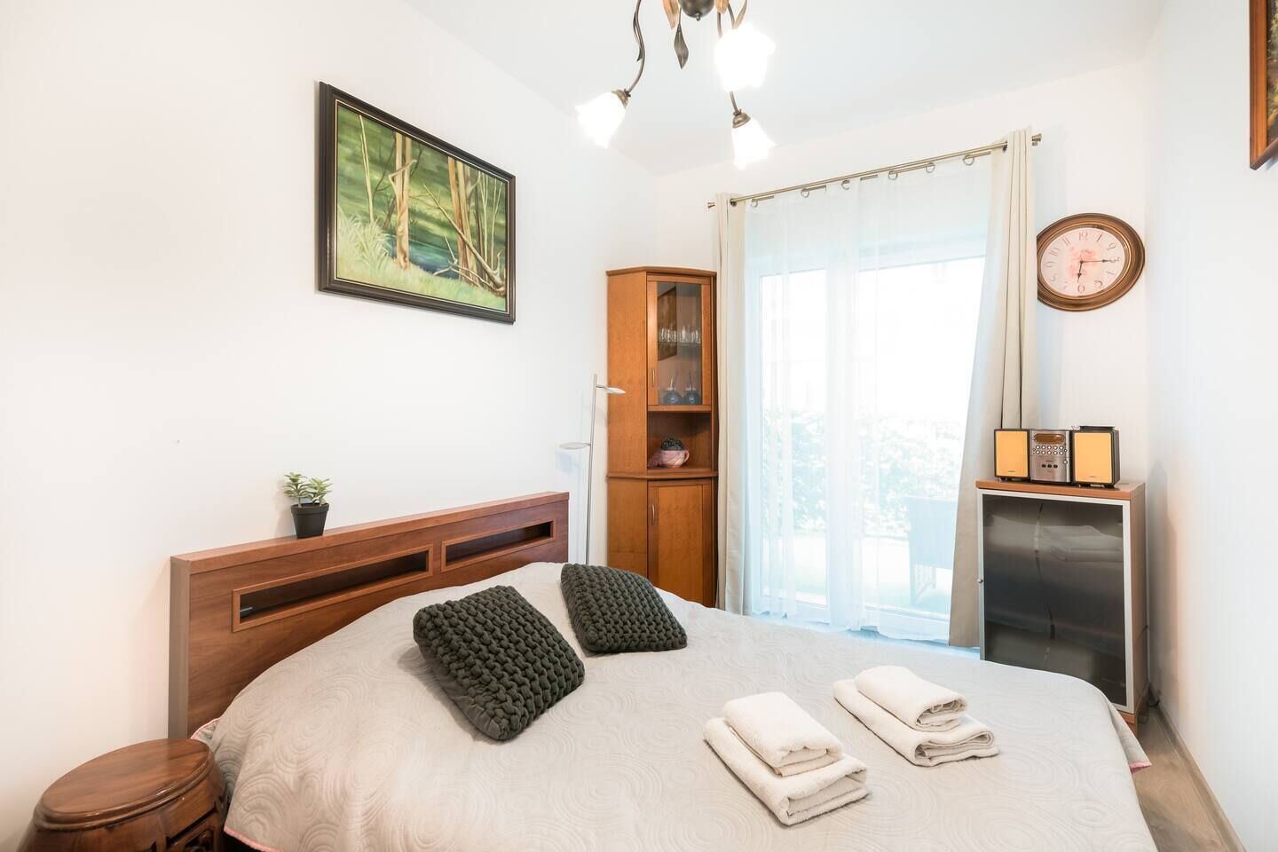 A bedroom with a bed, a wooden headboard, a cabinet, a bedside table with a radio, a window with curtains, and a painting on the wall.