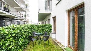 Un cortile con tavolo e sedie, circondato da un siepe e un edificio con ampie finestre.