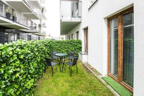 Ein Innenhof mit Tisch und Stühlen, umgeben von einer Hecke und einem Gebäude mit großen Fenstern.