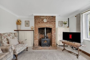 A living room with a brick fireplace, a floral sofa, a television, and a white cabinet. - Charming Suffolk Cottage, 3 Bedroom + Parking (Blyford)