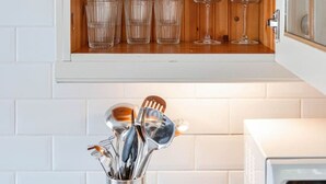 Een keuken met een houten kastplank met witte bekers en glazen, een metalen houder voor keukengerei, en een magnetron.