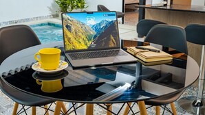 A laptop on a glass table with a yellow cup, a notebook, and a pen.