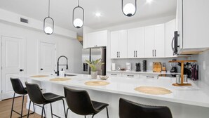 A modern kitchen with white cabinets, a central island with bar stools, pendant lighting, and a coffee machine. - Stylish Downtown Retreat Near the Alamodome (San Antonio)