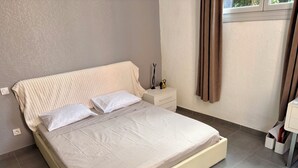 A neatly made bed with a white comforter and pillows, a bedside table with a lamp and a small decorative item, and a window with curtains. - Apartment T2/3 Corte center (Corte)