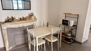 A dining area with a white table and chairs, a fireplace with Christmas decorations, a metal shelf with kitchen appliances, and a wall-mounted mirror. - Apartment T2/3 Corte center (Corte)