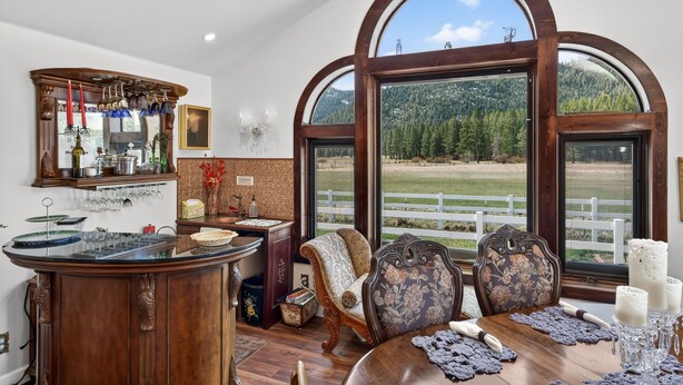 A dining area with a wooden table, chairs, and a view of a fenced field through large windows. - Mountain View Escape | Hot Tub, Fire Pit & Games (Sattley)