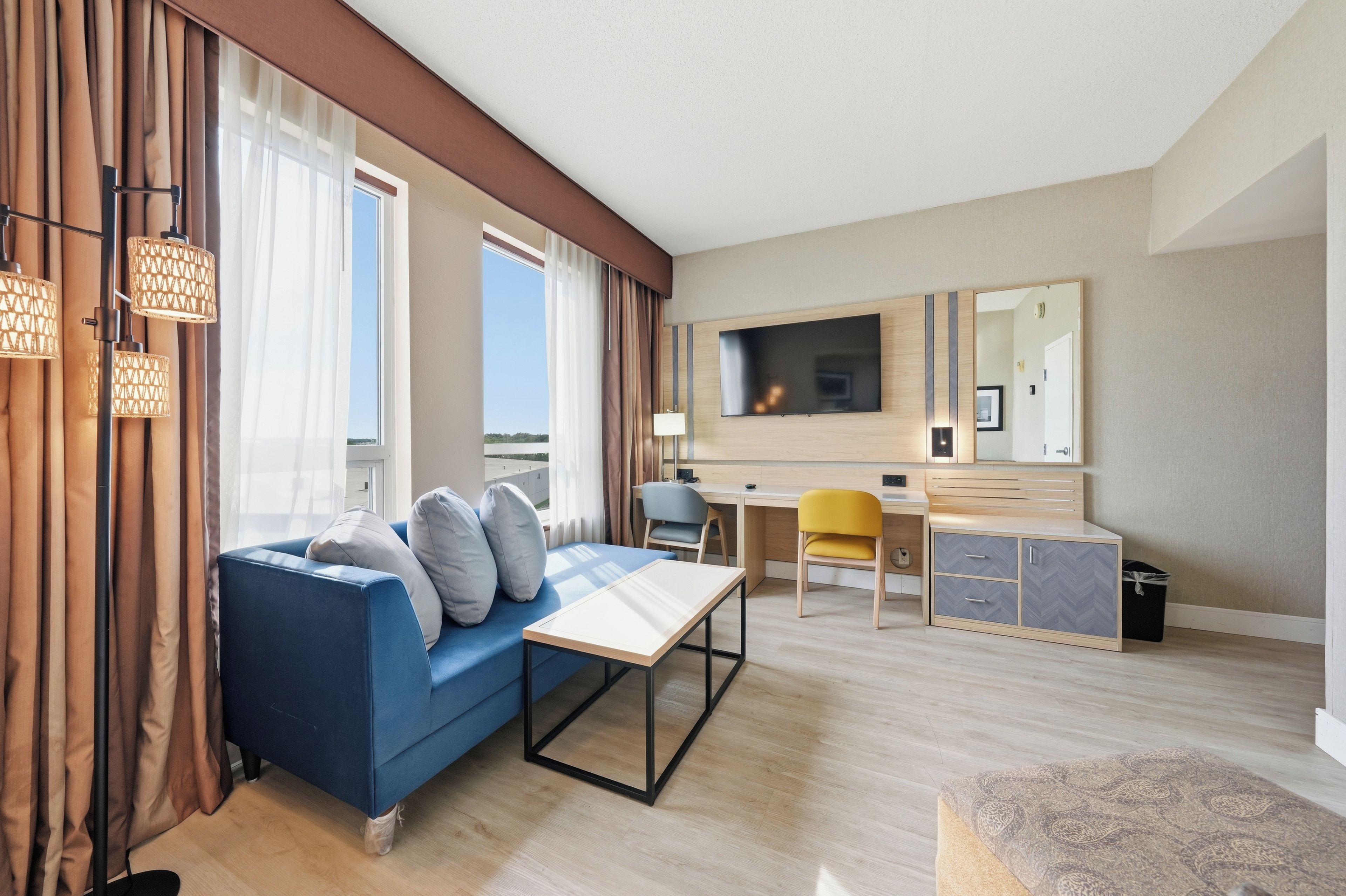 A modern hotel room with a blue sofa, a desk, a chair, a TV, and a large window with curtains.
