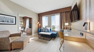 A hotel room with a sofa, a blue armchair, a small table, a TV mounted on the wall, and a window with curtains.