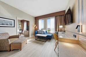 Une chambre d’hôtel avec un canapé, un fauteuil bleu, une petite table, un téléviseur fixé au mur et une fenêtre avec des rideaux.