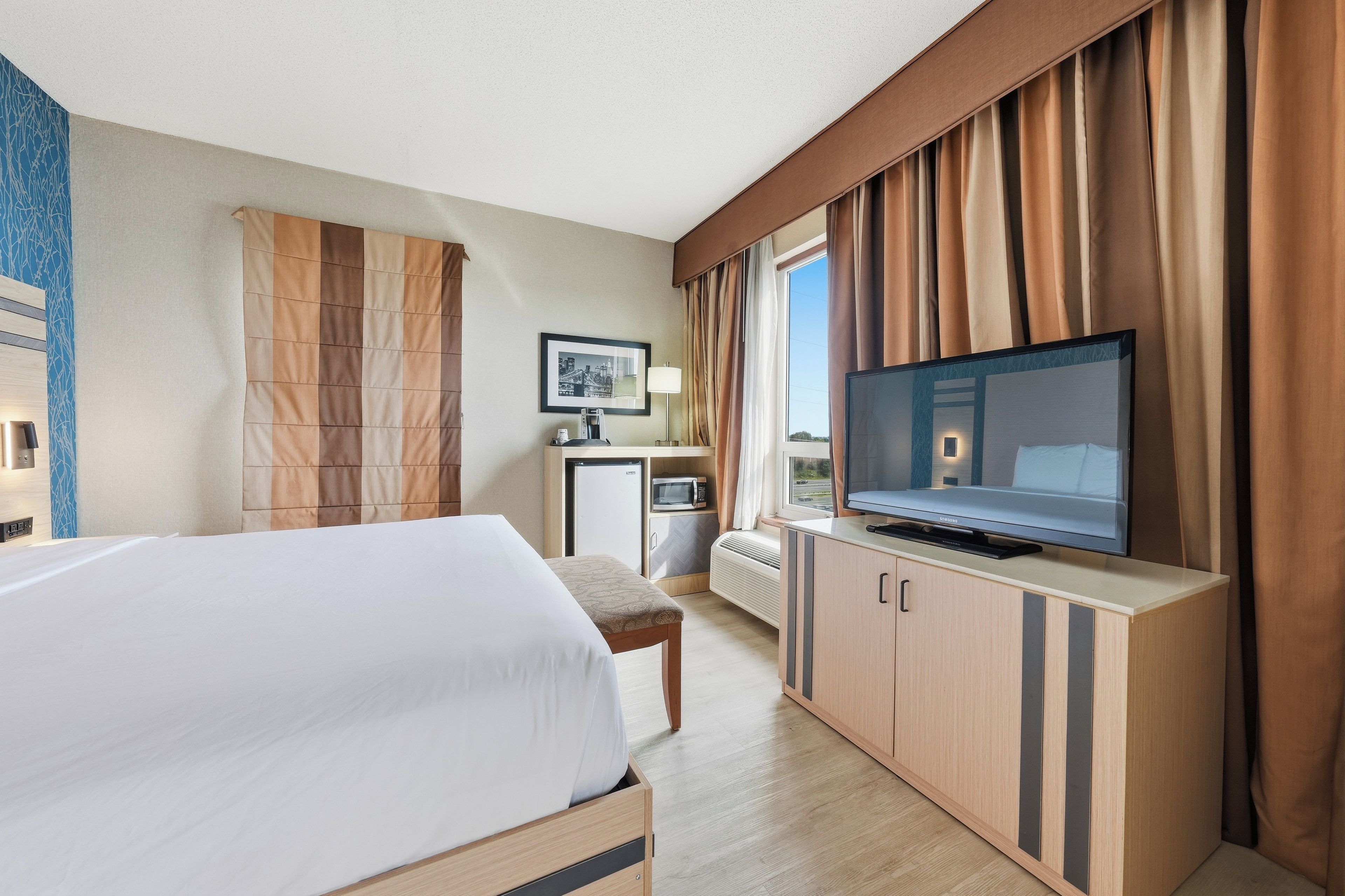 Habitación de hotel con una cama, televisor, escritorio con computadora y ventana con cortinas.