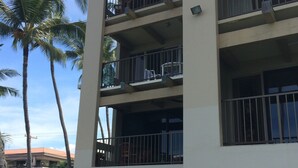 Un edificio a più piani con balconi, un'auto parcheggiata in un'area di parcheggio coperta e palme sullo sfondo.
