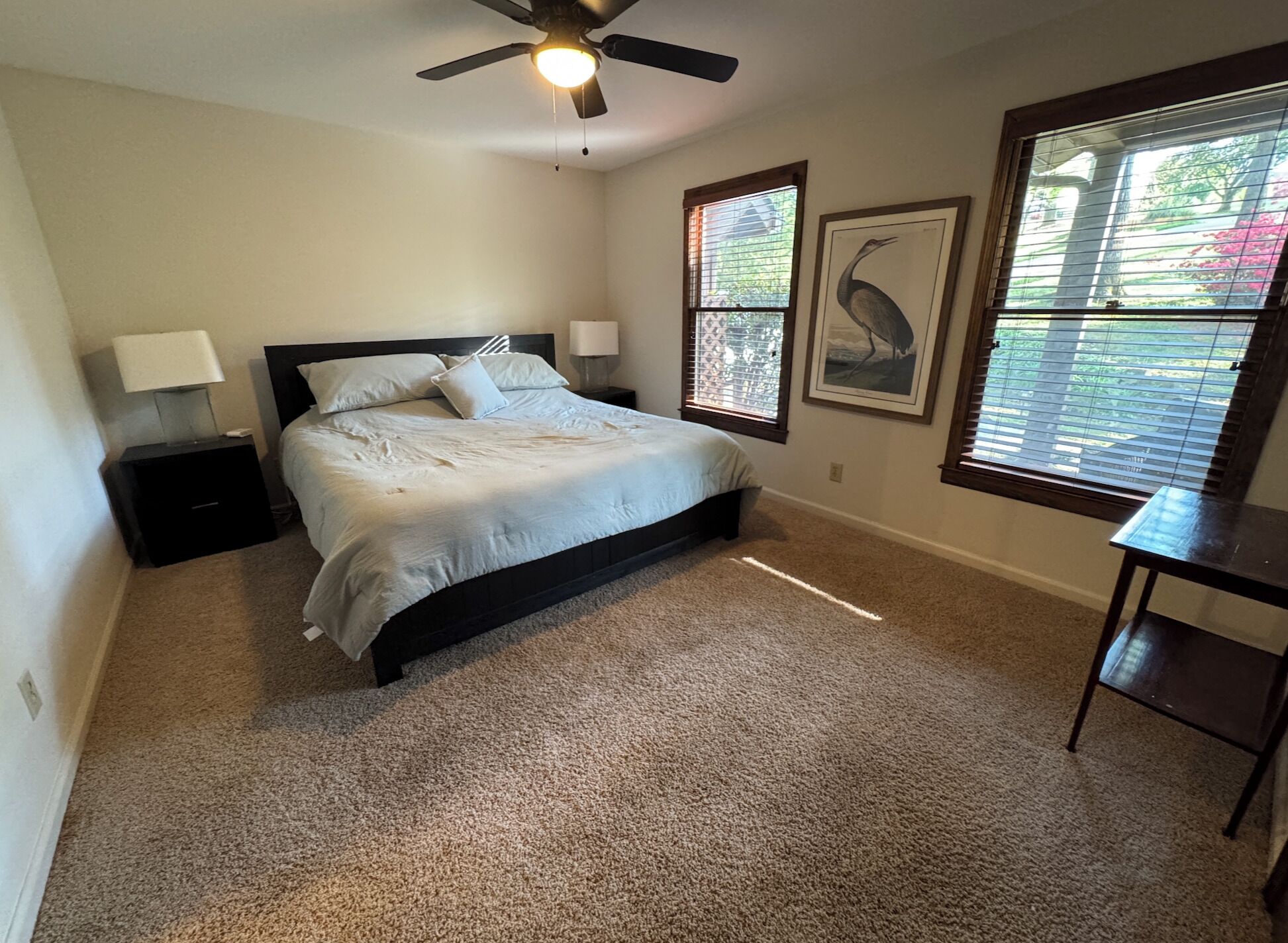 A bedroom with a bed, two bedside tables, a ceiling fan, and two windows with blinds.