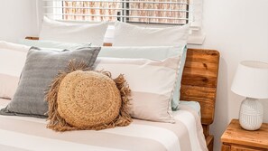 Una cama con varias almohadas, un cabecero de madera, una mesita de noche con una lámpara y una ventana con persianas.