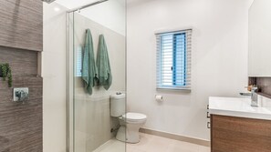 Un baño moderno con cabina de ducha de vidrio, inodoro, ventana con persianas y tocador de madera.